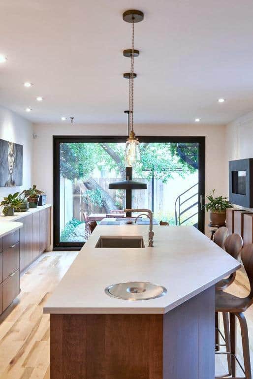 Cuisine moderne avec armoires blanches, touches de bois et un grand îlot avec luminaires suspendus et tabourets de bar. Un coin repas avec une table et des chaises en bois se trouve à côté d'un tableau, de plantes et d'appareils de cuisine sur le comptoir. Derrière l'ilot, une salle ç manger avec un magnifique foyer