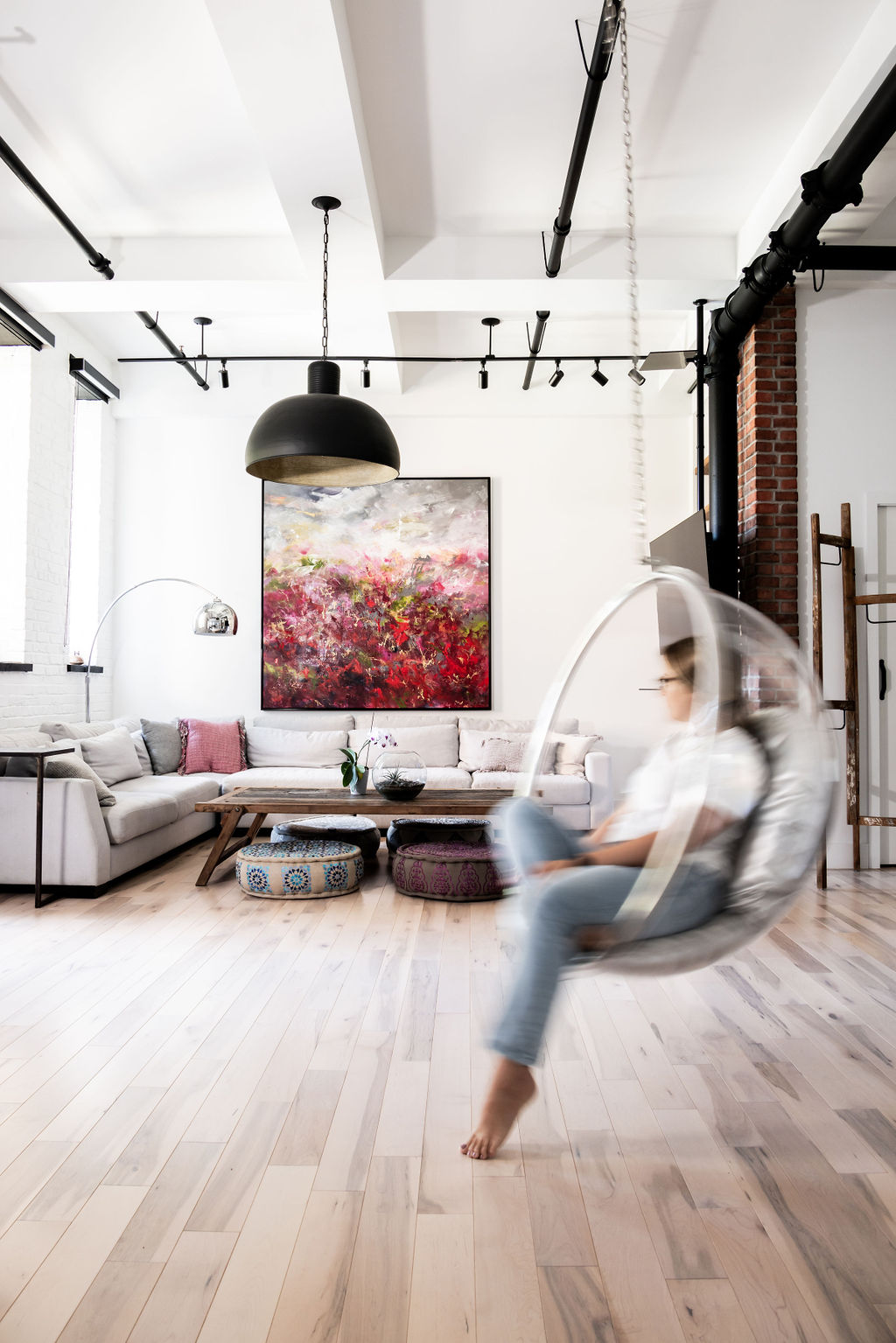 Un salon moderne et lumineux avec un canapé d'angle blanc, une peinture abstraite colorée et des coussins de sol ronds. Une personne est assise sur une chaise à bulles transparente suspendue au plafond. Des luminaires industriels sont visibles.
