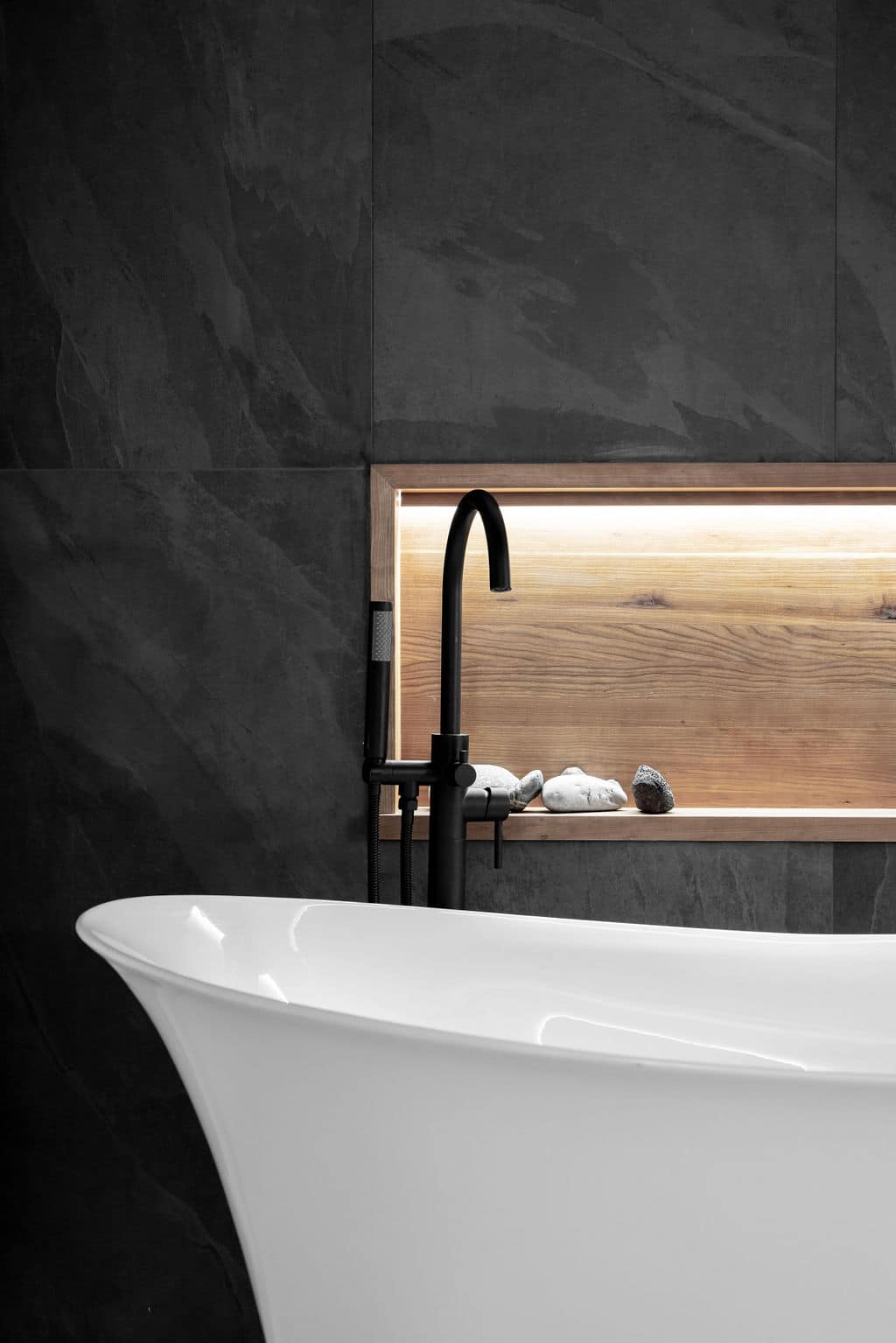 Une salle de bains moderne avec une baignoire blanche épurée au premier plan. Derrière la baignoire, il y a un mur avec une étagère en bois avec un éclairage d'ambiance et des pierres décoratives. Le mur est recouvert de carreaux d'ardoise foncée et un robinet noir mat est visible.